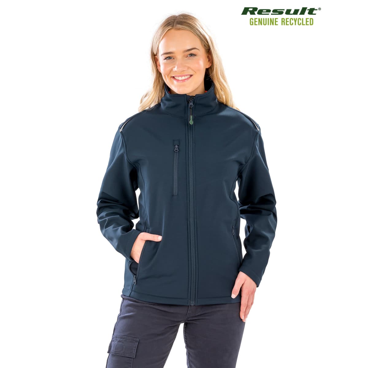 A person wearing a dark blue jacket with zipper pockets is smiling and standing with hands in pockets. The jacket is branded with Result Genuine Recycled. The background is plain white.