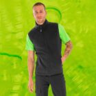 A man with short hair, wearing a bright green T-shirt and a black vest, stands against a vibrant lime green background. His right arm has tattoos, and he is looking towards the camera with a slight smile.