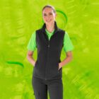 A woman wearing a black vest and bright green shirt poses with her hands in her pockets against a vibrant green background. She is smiling confidently.