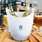 White ice bucket with three beer bottles in it, placed on a wooden table. Two glasses of beer on either side. A logo with a gear symbol and COMPANY text is on the bucket. Beer kegs are blurred in the background.