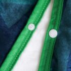 Close-up of a white button sewn onto fabric, with a green hem and blue-green patterned textile. The button is secured with a snap fastener. The fabrics appear soft and plush, suggesting a cozy garment or blanket.