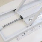 Close-up of a white mechanical device with a metal rod and LED strip. Components are arranged on a flat surface, including a lever and rectangular elements. The LED strip has multiple round lights and visible electrical connections.