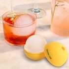 A glass of a red cocktail with a large ice cube sits beside a glass of a light pink drink with a straw. In the foreground is a yellow capsule labeled Drone Cam, opened to reveal a white sphere inside.