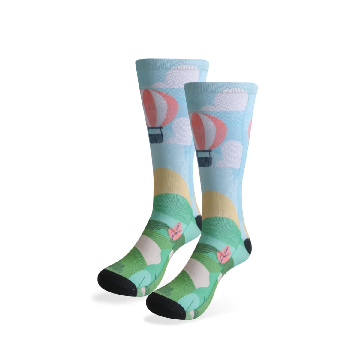 A pair of colorful socks featuring a whimsical scene with red and white hot air balloons floating among clouds above a green landscape, with plants and flowers at the bottom. The socks have black toes and heels.