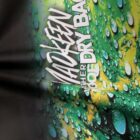 Close-up of a fabric with green and yellow hues resembling droplets on a surface. White text reads MADKEEN followed by THE DRY BAG along the folds of the material.