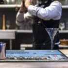 Bartender shaking drink behind a bar with an empty martini glass in the foreground.