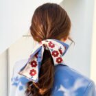 A woman with long brown hair is wearing a blue blazer. Her hair is tied in a low ponytail with a large, folded scarf featuring red flowers and blue edges. The background is a soft white wall.