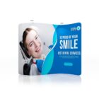 A woman at the dentist smiles while leaning back in a dental chair. Next to her, a dental professional is visible. The backdrop features text: Be Proud of Your Smile - Best Dental Services, with a logo and some placeholder text below.