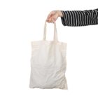 A hand in a black and white striped sleeve holds a plain, white tote bag against a white background.