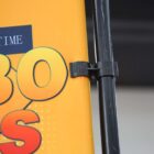 Close-up of a yellow and orange sign attached to a black pole with a plastic clip. Partial text visible, showing 30 in large, bold letters, along with other partially obscured elements.
