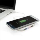 A smartphone is charging wirelessly on a charging pad next to an open laptop. The phone screen displays a green battery icon indicating charging. The laptop is connected to the charging pad via a USB cable.