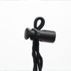 A close-up of a black plastic spring cord lock holding a looped black drawstring. The cord lock is cylindrical with a button on top, set against a plain white background.