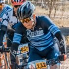 Three cyclists wearing helmets and sunglasses ride mountain bikes on a dirt path during a race. They are focused and wearing numbered race bibs. One cyclist in the foreground wears a blue kit with Ruteam on the front. Trees are blurred in the background.