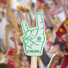 A person holds up a foam hand with the index and pinky fingers raised in a rock gesture. Cheer is printed on it. A cheering crowd is blurred in the background.