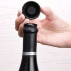 A persons hand is holding a black bottle cap above the neck of a wine bottle. The hidden label suggests the wine type is Moscato d’Asti. The background is a textured white wall.