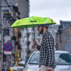 Man walking on a city street holding a green umbrella with a swirl design and escale logo. He wears a checkered shirt and beanie. Overcast sky and cars are visible in the background.