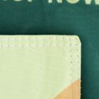 Close-up of a fabric with two layers. The background layer is dark green, partially displaying the text NOW. The foreground layer is a rectangular light green piece with visible white stitching around the edges.