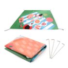 A green picnic blanket with a colorful design featuring food and musical items, laid out flat. Below it, the blanket is folded neatly with four metal stakes beside it. The design includes a guitar, pizza, grapes, and drinks.