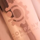 A metallic surface features an engraved logo resembling a gear with a dot and a curved line inside. Below is the text COMPANY with a placeholder for a slogan reading YOUR SLOGAN HERE. The surface has a glossy, reflective finish.