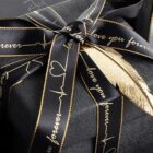 A black gift box wrapped with a ribbon featuring the words love you forever in gold script. A gold feather-shaped decoration is attached, enhancing the elegant presentation.