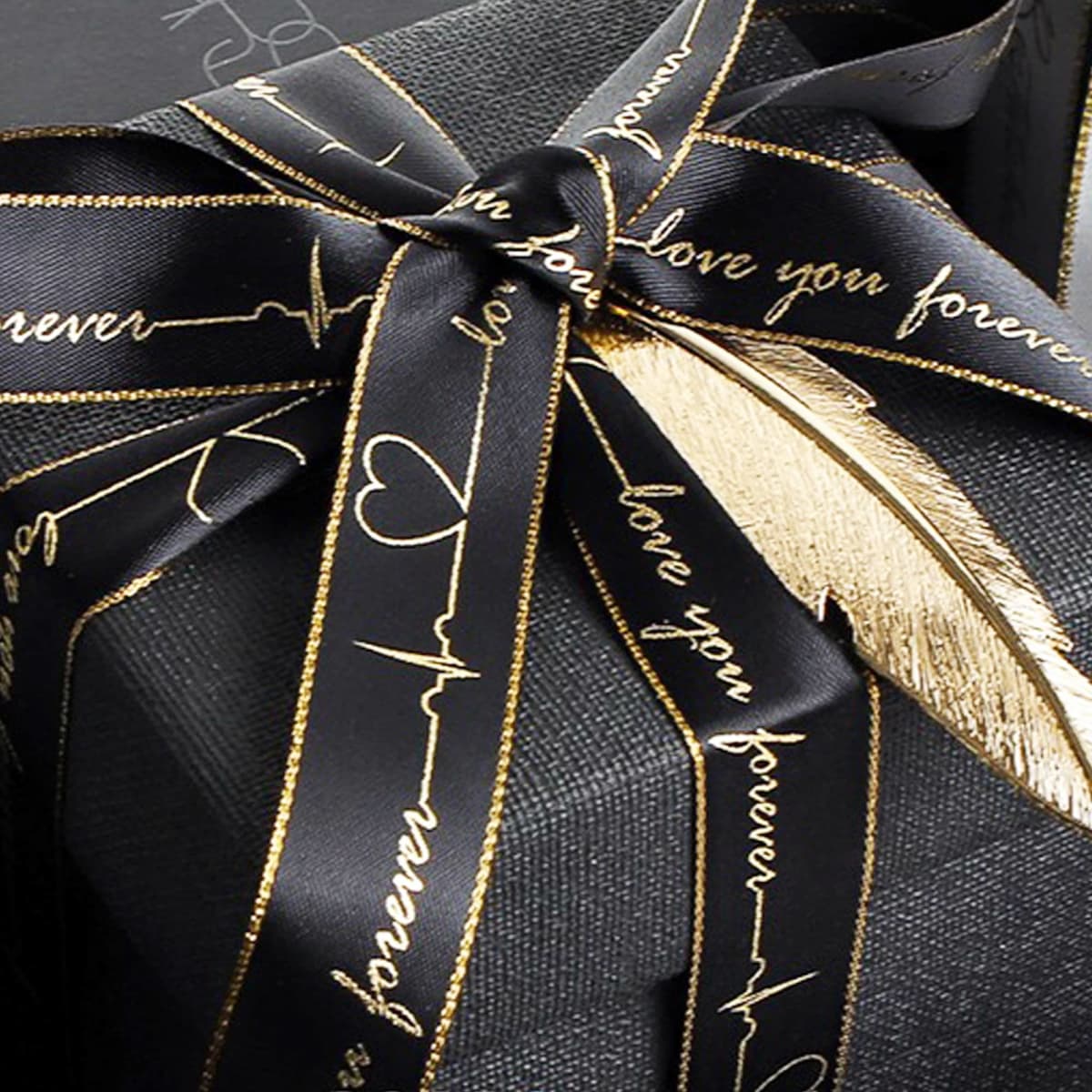 A black gift box wrapped with a ribbon featuring the words love you forever in gold script. A gold feather-shaped decoration is attached, enhancing the elegant presentation.