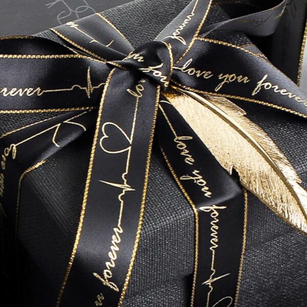 A black gift box adorned with a silky black ribbon featuring a gold script that reads love you forever. A gold feather accent is tucked under the bow, adding a luxurious touch to the presentation.
