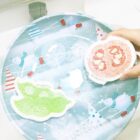 A person holding two fruit-themed scrub sponges—one green shaped like a lime and one red shaped like a grapefruit—above a decorative plate with a winter pattern featuring sleds and snowflakes.