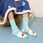 Person wearing mint green socks with snowflakes and snowman design, sitting on a blue floor. They have a blue blanket with white and green polka dots, featuring a white trim. Their feet rest on a yellow cushion.