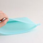 A hand holds a flexible, light blue silicone food storage bag, demonstrating its pliability. The bag has a semi-transparent appearance and a tab on one end for sealing. The background is plain and neutral.