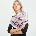 A woman with blonde hair wearing a pink patterned silk scarf draped over her shoulders, paired with a black top. The background is plain white.