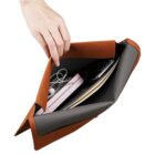 A hand holds an open brown pouch containing various items, including a smartphone, a pink book, earbuds, and a beige tablet. The pouch is partially unzipped, revealing the contents inside.