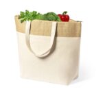A beige and brown tote bag filled with fresh vegetables, including a bunch of green parsley, a head of broccoli, and a red bell pepper, is shown against a white background.