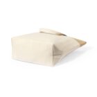 A beige canvas tote bag is shown from the side, laying on its flat base. It has a minimalist design with a single handle on top, and the bags fabric is slightly textured. The background is plain white.