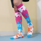 Person wearing colorful knee-high socks with a splash pattern and the words MAGIC MILK on one sock. The socks feature pink, white, blue, and orange colors. The person stands in a room with a light blue floor and beige wall.