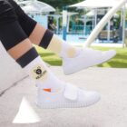 A person wearing black pants, white sneakers with Velcro straps, and white socks featuring a bear graphic and the text KIDSTORY is sitting outdoors on a sunny day with blurred umbrellas and greenery in the background.