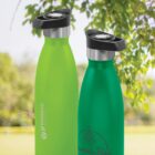 Two green reusable water bottles with black lids, outdoors near leafy branches.