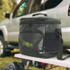 A black insulated cooler bag with Olive and Co written on it sits on a table outdoors. A silver SUV is parked in the background, and trees are visible. The cooler has a zippered top, front pocket, and a side mesh pocket.