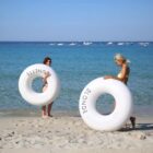 Two women on a sandy beach hold large white inflatable rings. The woman on the left wears a black bikini and holds a ring labeled BRUNETTE. The woman on the right wears a yellow bikini and holds a ring labeled BLONDE. The sea and boats are in the background.