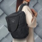 A person wearing a beige coat is carrying a black Osprey backpack on their shoulder. The background features a geometric gray wall pattern.
