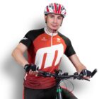 A person wearing a red and black cycling jersey and a red and white helmet is holding the handlebars of a bicycle, posing against a plain white background. They are also wearing fingerless cycling gloves.