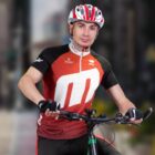 A person wearing a red and black cycling jersey and a white helmet is riding a bicycle. The background is blurred, suggesting movement.