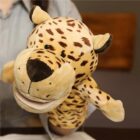 A person is holding a plush leopard hand puppet. The puppet has a smiling expression, with spotted fur, round ears, and a large snout. The background is indoors with a blurred wall.