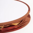 Close-up of a circular foldable item with a white top and brown edge. The bottom part is partially folded, revealing layers of fabric in earth tones. The background is plain and light-colored.