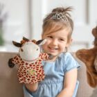 A young girl with a tiara sits on a couch, smiling. She is holding a giraffe puppet with brown spots and wearing a blue dress. The room has a softly blurred background with light-colored walls.