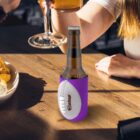 A person holding a glass of beer and clinking it with another beer bottle wrapped in a purple, football-shaped koozie on a wooden table. A bowl of chips is visible on the table.