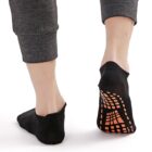 A person wearing black no-show socks with orange rubber grips on the soles, walking on a white surface. They wear dark gray pants, and the image focuses on their lower legs and feet from behind.