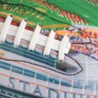 Close-up of a football with a colorful design. The surface features laces and a painted image of a stadium with green fields and small figures, likely representing a crowd or players, around it.