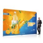 A large advertising banner displays a bottle of orange juice with splashes and sliced oranges. A man in a suit stands next to it, smiling. The banner showcases vibrant orange and blue colors, emphasizing the freshness and natural ingredients of the juice.