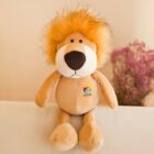 A plush lion toy with a fluffy orange mane is seated on a surface. It features a logo on its chest with the word ACTIVE. In the background, there is a bouquet of small pink and white flowers on a beige backdrop.