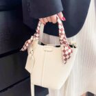Person holding a small beige handbag with checkered scarf wrapped around the handles, wearing a dark jacket and white pleated skirt. The bag has a rectangular tag attached. The person has red nails.